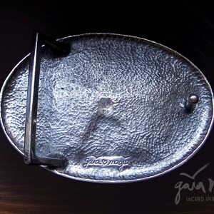 May include: A silver oval belt buckle with a textured surface and a small metal loop on the side. The buckle is engraved with the words "Gaia Magia" and a heart symbol.