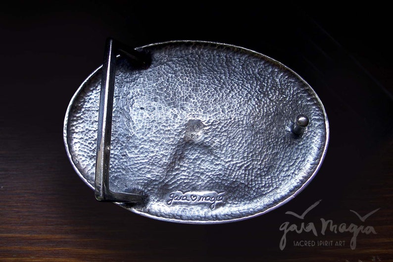 May include: A silver oval belt buckle with a textured surface and a small metal loop on the side. The buckle is engraved with the words "Gaia Magia" and a heart symbol.