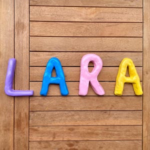 Puede incluir: Letras coloridas y hechas a mano que deletrean el nombre "Clara". Las letras son de varios colores brillantes: naranja, morado, azul, rosa y amarillo. Dispuestas sobre una superficie de madera.