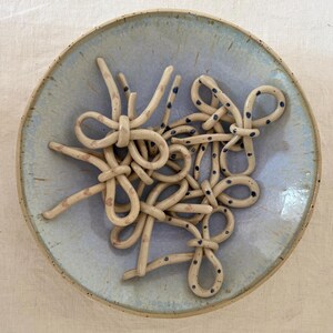 Op de afbeelding: Een handgemaakte keramische kom met een lichtblauwe glazuur. De kom bevat een beige, geknoopte keramische sculptuur met zwarte stippen. De rand van de kom is een donkerdere tint blauw en de achtergrond is neutraal.