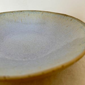 Pode incluir: Um prato de cerâmica feito à mão com um centro cinza-azulado claro e uma borda marrom quente. O prato tem uma superfície ligeiramente texturizada e um design simples e elegante. Perfeito para servir comida ou como peça decorativa.