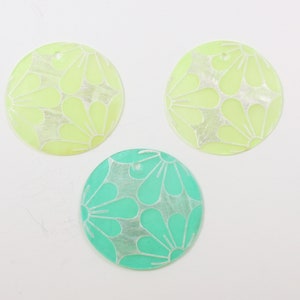 May include: Three round, decorative, iridescent, plastic embellishments. One is green with a white floral pattern, and two are yellow with a white floral pattern.