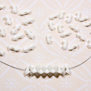 May include: A close-up of white ceramic beads strung on a silver wire. The beads are shaped like a pointed arrowhead and are arranged in a repeating pattern. There are also loose beads scattered around the wire.