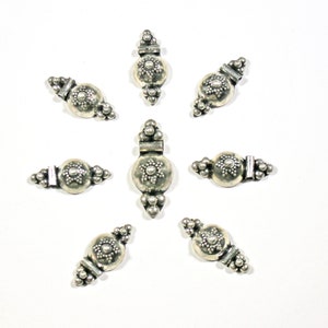 May include: Eight silver metal beads with a decorative design. Each bead has a round center with a raised star-shaped pattern and is surrounded by small beads. The beads are arranged in a star shape.