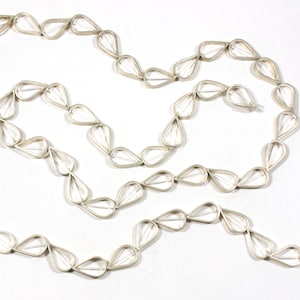 Op de afbeelding: Een zilveren ketting gemaakt van gekoppelde traanvormige stukken. De ketting ligt op een wit oppervlak.