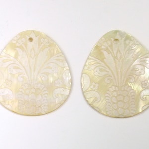 May include: Two white oval-shaped shell pendants with a white floral design. The pendants have a hole at the top for stringing.