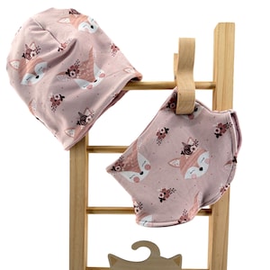 Könnte beinhalten: Eine altrosa Mütze und ein passendes Halstuch mit einem wiederkehrenden Muster aus Füchsen mit Blumenkränzen. Die Artikel sind auf einer Leiter aus hellem Holz präsentiert, an der auch ein Holzkleiderbügel in Fuchskopfform mit dem Schriftzug Hanna Bell hängt.