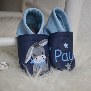 Könnte beinhalten: Ein Paar blaue Babyschuhe mit hellblauer Innenausstattung. Ein Schuh hat einen gestickten Esel, der andere Schuh hat den Namen "Paul" gestickt.