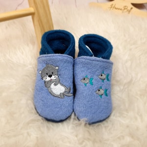 Könnte beinhalten: Ein Paar blaue Babyschuhe mit einem gestickten Otter- und Fischdesign. Die Schuhe haben ein dunkelblaues Fleecefutter.