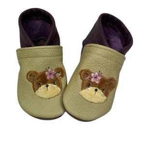 Könnte beinhalten: Ein Paar hellgrüne Lederschuhe für Babys mit gestickten braunen Bärenköpfen auf der Vorderseite. Die Schuhe haben ein violettes Futter und sind aus weichem Leder.