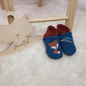 Könnte beinhalten: Blaue und braune Filz-Babyschuhe mit einem Fuchs-Design und Stickereien von Federn auf der Vorderseite. Die Schuhe liegen auf einer weißen, flauschigen Oberfläche.