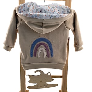 Könnte beinhalten: Beigefarbener Kapuzenpullover mit Regenbogen-Applikation auf dem Rücken. Das Kapuzenfutter hat ein Blumen- und Regenbogenmuster. Der Hoodie wird auf einer Holzleiter mit einem fuchsformigen Schild mit der Aufschrift "Hanna Bell" präsentiert.