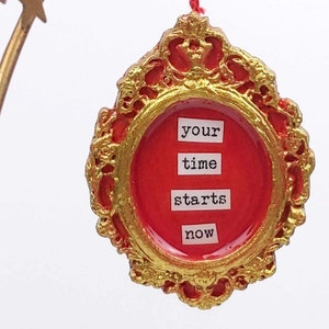 Może przedstawiać: Ozdoba dekoracyjna z czerwonym środkiem i złotą, ozdobną ramą. Tekst "your time starts now" jest wyświetlany białymi literami. Ozdoba zawieszona jest na czerwonym sznurku.