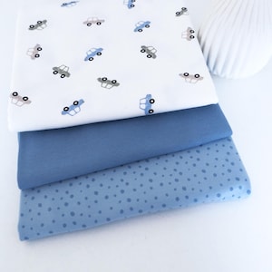 Può includere: Tre pezzi di tessuto piegati in diverse tonalità di blu. Lo strato superiore è bianco con un motivo di piccole auto blu e grigie. Lo strato intermedio è blu medio unito e lo strato inferiore è azzurro con un motivo a pois.