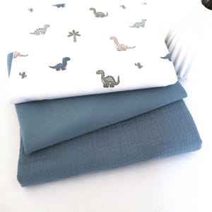 Könnte beinhalten: Drei gefaltete Stoffstücke, die übereinander liegen. Das oberste Stück ist weiß mit einem sich wiederholenden Muster aus kleinen Dinosauriern und Kakteen in Blau-, Grün- und Beigetönen. Die mittleren und unteren Stücke sind aus einfarbigem, strukturiertem blauem Stoff.