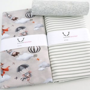 Könnte beinhalten: Stoffpakete mit einem skurrilen Druck von Zeichentricktieren in Heißluftballons und Fallschirmen sowie einem Streifenmuster. Das Material ist mit "stoffrevier Jersey" gekennzeichnet. Ein gerolltes Stück grauer Stoff ist ebenfalls vorhanden.