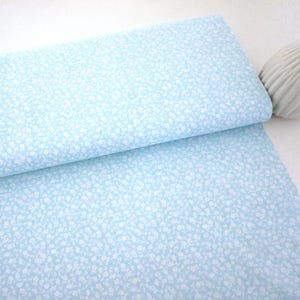 Può includere: Un rotolo di tessuto azzurro con un delicato motivo floreale bianco. Il tessuto è piegato, rivelando il motivo e la trama. Lo sfondo è bianco.