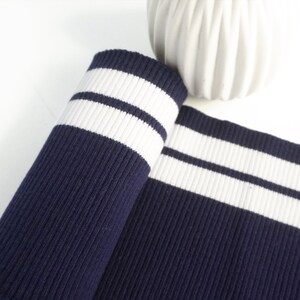 Könnte beinhalten: Gerollter marineblauer Stoff mit zwei weißen Streifen. Die gerippte Textur des Stoffes ist sichtbar. Der Stoff wird wahrscheinlich für Kleidung oder Accessoires verwendet.