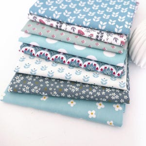 May include: A stack of seven folded fabric pieces featuring various patterns in shades of blue, white, and grey. The designs include white leaf motifs, floral prints, large white polka dots, and small white daisy-like flowers on solid backgrounds.