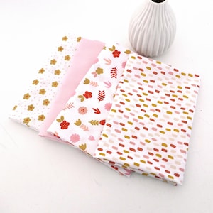 Könnte beinhalten: Vier gefaltete Stoffstücke in Weiß, Rosa und Mustern. Ein Stoff hat kleine goldene Blumen, ein anderer rote und goldene Blumenmuster und der letzte ein gepunktetes Muster in Rosa, Gold und Rot. Im Hintergrund steht eine weiße Vase.