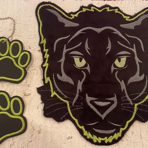 Könnte beinhalten: Schwarze Panthergesicht- und Pfotenabdruck-Patches mit neongrünen und grauen Akzenten. Ein Pfotenabdruck-Patch hat eine silberne Kette. Das Panthergesicht und die Pfotenabdrücke sind auf schwarzem Hintergrund gestickt.