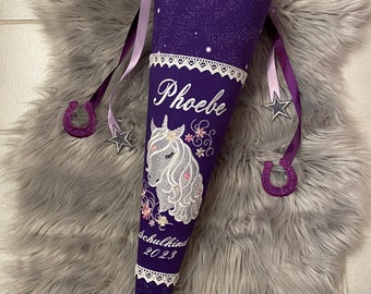 May include: A purple school cone with a white lace trim and a starry pattern. It features a white embroidered unicorn, the name "Phoebe", and the text "Schulkind 2023". The cone is decorated with ribbons, stars, and a horseshoe.