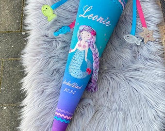 Pode incluir: Um cone escolar com tema de sereia em tons de azul e roxo, com o nome "Leonie" bordado na frente. O cone apresenta uma ilustração de sereia, acabamentos decorativos e uma cauda roxa. Inclui enfeites em forma de peixe.