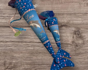 Puede incluir: Dos conos escolares azules y blancos con ilustraciones de delfines, estrellas rosas y ribetes ondulados. Las bolsas tienen un diseño de cola de sirena y el nombre "Lea" está impreso en ellas. Una bolsa tiene el texto "Schulkind 2025".