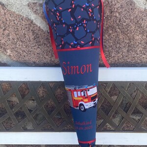 Könnte beinhalten: Eine marineblaue Schultüte mit rotem und blauem Band. Der Kegel hat ein Muster aus roten und blauen Formen. Der Name "Simon" ist rot aufgedruckt, zusammen mit einer Feuerwehrwagen-Grafik und dem Text "Schulkind 16.09.2023."