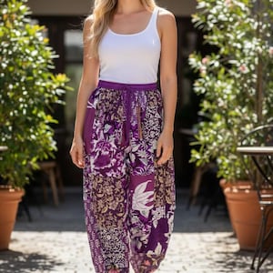 May include: A person wearing a white vest top and purple patterned trousers. The trousers have a floral design in shades of purple, white, and brown. The trousers have a drawstring waist and are paired with brown sandals. The background includes potted plants and a brick walkway.