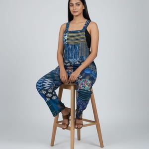 Può includere: Una donna indossa una tuta blu fantasia con un top nero sotto, seduta su uno sgabello di legno. La tuta presenta vari motivi blu. Lo sgabello è in legno chiaro.