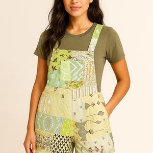 Op de afbeelding: Een patchwork romper in groen- en geeltinten, met verschillende patronen en texturen. Het kledingstuk in shorts-stijl heeft zakken en verstelbare bandjes met houten knopen. Het model draagt er een groen t-shirt onder.
