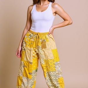 Peut inclure: Pantalon jaune en patchwork avec taille élastique et cordon de serrage. Le pantalon présente divers carrés de tissu à motifs dans les tons jaune et crème. Porté avec un débardeur blanc.