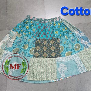 Op de afbeelding: Een patchwork katoenen rok met diverse bloemen- en geometrische patronen in blauwe, groene en gele tinten. De rok heeft een elastische tailleband en een uitlopend silhouet. Het woord "Cotton" is in het blauw geschreven.