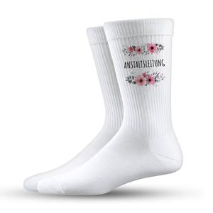Könnte beinhalten: Ein Paar weiße Socken mit dem schwarzen Schriftzug "ANSTALTSLEITUNG", umrahmt von einem Blumendesign mit rosa Blüten und grünen Blättern. Die Socken sind gerippt und scheinen aus einem weichen Material zu sein.