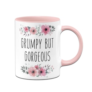 Könnte beinhalten: Weiße Keramik-Tasse mit rosa Innenraum und Henkel. Die Tasse zeigt den Text "GRUMPY BUT GORGEOUS" in Schwarz, mit floralen Akzenten in Rosa und Grün. Eine charmante und humorvolle Geschenkidee.