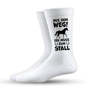 Könnte beinhalten: Ein Paar weiße Socken mit schwarzem Text und einer Pferdezeichnung. Der Text lautet "AUS DEM WEG! ICH MUSS ZUM STALL", was übersetzt "Aus dem Weg! Ich muss in den Stall" bedeutet. Die Socken haben eine gerippte Textur.