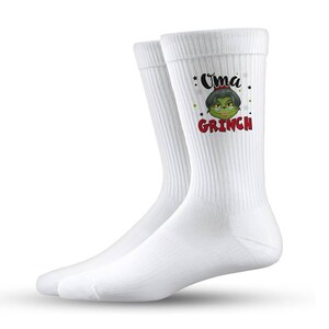 Könnte beinhalten: Weiße Crew-Socken mit dem Text "Oma Grinch" und einem Cartoon-Bild einer grünen Figur. Die Socken haben eine gerippte Textur und sind aus einem weichen Material gefertigt.