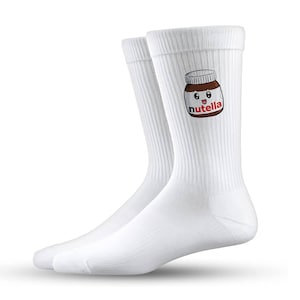 Könnte beinhalten: Ein Paar weiße Crew-Socken mit einem Nutella-Glas-Motiv. Das Glas ist braun mit einem weißen Etikett und dem Wort "nutella" in Rot. Die Socken sind gerippt und haben einen lässigen Stil.