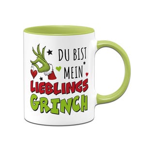 Könnte beinhalten: Weiße Keramik-Tasse mit grünem Innenraum und Griff. Die Tasse zeigt ein Grinch-Motiv mit dem Text "DU BIST MEIN LIEBLINGS GRINCH" in Rot und Grün, zusammen mit einer Grinch-Hand, die ein Herz hält.