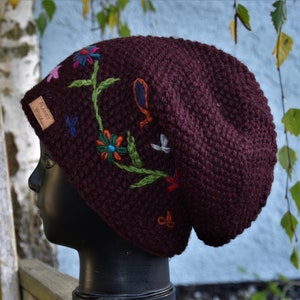 Könnte beinhalten: Eine burgunderfarbene, schlaffe Mütze mit bunten, gestickten Blumen und Blättern. Die Mütze hat ein kleines braunes Leder-Etikett mit dem Text "Forest Wool" darauf.