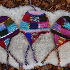 Könnte beinhalten: Drei bunte Strickmützen mit Ohrenklappen und langen geflochtenen Bändern. Die Mützen sind aus verschiedenenfarbigen Garnen gefertigt und haben ein Patchwork-Design.