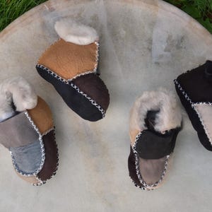 Könnte beinhalten: Vier Paar braune, graue und schwarze Lederschlappen mit weißem Fellfutter. Die Schlappen liegen auf einer hellbraunen Oberfläche.