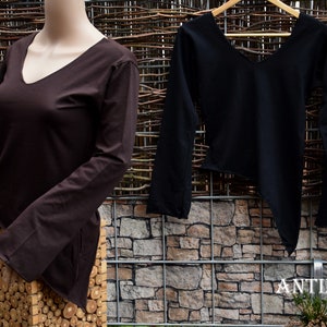 Könnte beinhalten: Ein dunkelbraunes Langarmshirt auf einer Schaufensterpuppe und ein schwarzes Langarmshirt mit asymmetrischem Saum an einem Holzkleiderbügel. Beide Shirts haben einen V-Ausschnitt und glatten Stoff. Der Text ANTIMJA ist unten rechts zu sehen.