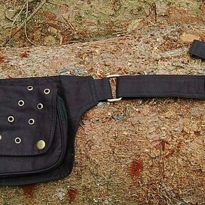 May include: A black canvas belt bag with a large pocket and multiple metal eyelets. The bag has a long adjustable strap with a silver buckle.