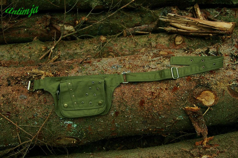 May include: A green canvas fanny pack with metal eyelets and a buckle closure. The pack is lying on a log in a forest setting.