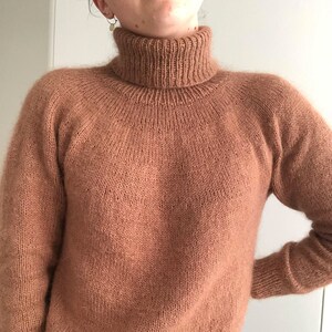 May include: A light brown knitted turtleneck jumper. The jumper features a high, ribbed collar and long sleeves. The knitwear appears soft and slightly fuzzy. The jumper is a solid colour and has a relaxed fit.