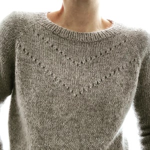 May include: A light grey knitted sweater with a crew neck and raglan sleeves. The sweater has a decorative openwork pattern across the chest. The knitwear has a textured appearance and a neutral colour.