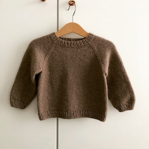 Könnte beinhalten: Ein brauner Strickpullover mit langen Ärmeln, der auf einem Holzbügel präsentiert wird. Der Pullover hat einen Rundhalsausschnitt und einen gerippten Saum und Bündchen. Das Kleidungsstück ist aus weichem, strukturiertem Garn gefertigt.