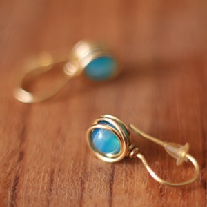 May include: A pair of gold wire earrings with blue gemstone accents. The earrings are made with a simple wire design and feature a round blue gemstone in the center.